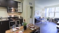 This single-floor residence offers a thoughtfully designed open-concept layout that seamlessly connects the kitchen, dining, and living areas into one cohesive living space. The modern kitchen is outfitted with dark cabinetry, stainless steel appliances including a gas range and built-in microwave, and light-toned countertops that provide a clean contrast. Upper cabinets with open shelving offer convenient storage and display space. Adjacent to the kitchen, the dining area features a warm wood table set for entertaining, complemented by elegant wine glasses and decorative greenery that add a welcoming touch. A discreet in-unit laundry closet with louvered doors is tucked neatly between the kitchen and living areas, adding practical functionality without interrupting the flow of the space. The living room is anchored by a tufted leather sofa and a sleek glass coffee table, resting on rich hardwood flooring that runs throughout the space. Large windows allow natural light to fill the room, creating a bright and airy ambiance. Wall art and carefully chosen accent pieces contribute to the space's modern yet comfortable character, making it both stylish and highly livable.