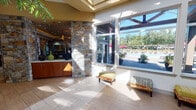 Located at 1302 NE Mount Constance Lane in Bremerton, Washington, this single-floor community clubhouse offers a comprehensive suite of amenities designed for both relaxation and active living. The welcoming entry features large floor-to-ceiling windows that flood the space with natural light and offer direct views of the outdoor pool area, framed by lush Pacific Northwest greenery. A prominent natural stone column anchors the entryway, setting an earthy, upscale tone carried throughout the facility. The main lounge is a generously sized gathering space with a coffered, tray ceiling accented by warm ambient lighting and dramatic sculptural pendant fixtures. Plush seating, built-in shelving, a wall-mounted television, and a reception or service counter make this an inviting hub for socializing. A reception area with a mosaic-tiled counter and warm wood cabinetry provides a polished point of contact. The fitness center is expansive and bright, featuring soaring vaulted ceilings, wall-to-wall mirrors, and large windows overlooking the exterior. It is outfitted with a full range of cardio and strength equipment, with a glass-walled partition separating additional workout zones. A dedicated racquetball court with hardwood floors, high ceilings, and glass panel access doors rounds out the athletic offerings. Carpeted corridors with frosted glass doors connect the various wings seamlessly.