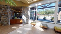 Located at 1302 NE Mount Constance Lane in Bremerton, Washington, this single-floor community clubhouse offers a comprehensive suite of amenities designed for both relaxation and active living. The welcoming entry features large floor-to-ceiling windows that flood the space with natural light and offer direct views of the outdoor pool area, framed by lush Pacific Northwest greenery. A prominent natural stone column anchors the entryway, setting an earthy, upscale tone carried throughout the facility. The main lounge is a generously sized gathering space with a coffered, tray ceiling accented by warm ambient lighting and dramatic sculptural pendant fixtures. Plush seating, built-in shelving, a wall-mounted television, and a reception or service counter make this an inviting hub for socializing. A reception area with a mosaic-tiled counter and warm wood cabinetry provides a polished point of contact. The fitness center is expansive and bright, featuring soaring vaulted ceilings, wall-to-wall mirrors, and large windows overlooking the exterior. It is outfitted with a full range of cardio and strength equipment, with a glass-walled partition separating additional workout zones. A dedicated racquetball court with hardwood floors, high ceilings, and glass panel access doors rounds out the athletic offerings. Carpeted corridors with frosted glass doors connect the various wings seamlessly.