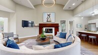 Located in Scottsdale, Arizona, this single-floor community space combines contemporary design with warm, inviting interiors throughout. The central lounge area is an immediate focal point, featuring soaring vaulted ceilings, a striking circular pendant light fixture, and a bold wood-paneled fireplace surround with a built-in firebox. A curved sectional sofa anchors the seating area atop a geometric area rug, creating an intimate gathering space ideal for socializing. Curated artwork, including a large equestrian portrait above the fireplace, adds character and sophistication.

Adjacent to the lounge, a sun-filled sitting nook lined with floor-to-ceiling windows offers panoramic views of the desert landscaping and surrounding community, bathing the space in abundant natural light. Light wood-tone flooring flows consistently throughout, unifying each area.

The kitchen is well-appointed with white upper cabinetry, stainless steel appliances, a rich wood-accented island with bar seating, and pendant lighting — making it functional for both casual use and events. A dedicated dining area features large windows framing lush outdoor views, while a separate formal dining space with a full-sized table provides versatility for meetings or gatherings. The overall ambiance is clean, modern, and welcoming, reflective of Scottsdale's upscale desert aesthetic.