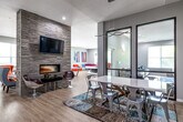 Located at 3306 Lincoln Way in Ames, Iowa, this single-floor community space offers a generously sized, thoughtfully designed amenity area built for both socializing and productivity. The open floor plan flows naturally between distinct zones, all finished with light wood-style flooring that unifies the layout and enhances brightness throughout.

The centerpiece of the main gathering area is a dramatic floor-to-ceiling stacked stone accent wall, home to a sleek linear fireplace and a wall-mounted flat-screen TV. Modern swivel chairs and lounge seating are arranged around the fireplace, creating an inviting focal point. Adjacent to this is a stylish dining area featuring a white table paired with mixed metal and modern chairs, illuminated by a contemporary pendant light fixture.

The living room area offers comfortable neutral sofas and armchairs accented with blue and patterned pillows, anchored by a bold blue abstract area rug. Black-framed glass partition doors add an architectural element while maintaining visual connectivity between spaces.

A dedicated conference room provides a focused work environment, equipped with a white meeting table, rolling chairs, a wall-mounted TV, and a whiteboard. A spacious game room with large windows allowing abundant natural light accommodates a ping pong table and foosball table. Colorful contemporary artwork is displayed throughout, contributing to the vibrant and energetic ambiance of the overall space.