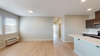 This single-floor unit offers a clean, contemporary design with consistent light wood-tone flooring and neutral warm-gray walls throughout, creating a cohesive and inviting atmosphere. The open-concept living area is generously proportioned, with recessed and flush-mount lighting illuminating the space. A peninsula counter connects the living area to the kitchen, providing a natural transition between the two zones. The kitchen is well-appointed with dark espresso-toned shaker-style cabinetry, crisp white countertops, and a full suite of stainless steel appliances including a refrigerator, range, and over-the-range microwave. A undermount sink is integrated into the peninsula, adding functionality. The bedroom features a large window fitted with horizontal blinds, allowing for ample natural light and outdoor views. A hallway connects the main living spaces to the private rooms, with a door providing access to the exterior. Two bathrooms are present: one featuring a soaking tub and shower combination with a large vanity mirror, and another with a walk-in shower stall, both outfitted with matching dark wood cabinetry and white countertops. The overall design is modern, practical, and well-finished throughout.