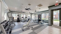 Located at 11212 Westpark Dr in Houston, TX, this fitness center offers a generously sized, single-floor layout designed to accommodate a full range of workout activities. The space is divided into two distinct zones: a carpeted section with a checkered gray tile pattern housing cardio equipment—including treadmills, ellipticals, and a cable machine—and a light hardwood flooring area dedicated to functional training, featuring spin bikes, kettlebells, a punching bag, a power rack, and open floor space for stretching or group exercise. Large windows line the perimeter walls, flooding the interior with natural light and offering views of the surrounding outdoor greenery. Multiple oversized ceiling fans are distributed throughout, ensuring consistent air circulation. Recessed lighting complements the bright, airy atmosphere. The neutral gray and white color palette gives the space a clean, modern aesthetic. A wall-mounted flat-screen TV is positioned near the entrance, alongside a dedicated stretching and mat area. Glass double doors provide a welcoming entry point. The overall design prioritizes both functionality and comfort, making it a comprehensive facility well-suited for a wide variety of fitness routines.