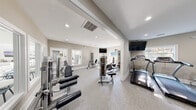 Located at 411 Gregory Dr in Cary, NC, this fitness center offers a well-appointed exercise space designed to support a full range of workout routines. The facility is laid out across a single floor with a functional, open floor plan that maximizes usability and flow between equipment areas.

The interior features neutral, warm-toned walls paired with light carpeted flooring, creating a clean and inviting atmosphere. Recessed lighting throughout ensures the space is consistently well-lit, while multiple windows allow natural light to filter in and provide views of the surrounding outdoors.

The cardio zone is equipped with treadmills, an elliptical trainer, and a stationary bike, all positioned beneath a wall-mounted flat-screen television for entertainment during workouts. The strength training area includes multiple cable and resistance machines, a weight bench, and a dedicated free weight section with a full dumbbell rack. A medicine ball storage tower offers a variety of weighted balls for functional fitness exercises.

Large wall-mounted mirrors along one section of the facility enhance the sense of space and allow users to monitor their form during exercise. The room also features a separate entry door providing direct outdoor access. The overall design is practical and modern, prioritizing comfort and functionality for an effective fitness experience.