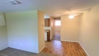 This single-story space offers a straightforward, functional layout with a clean and practical design. The main living area features warm hardwood-style flooring and neutral-toned walls that create a bright, open atmosphere. A ceiling fan is centrally mounted, providing both ventilation and ambient lighting to the space.

The layout flows seamlessly from the living area toward a compact kitchen, which is partially visible and includes wood-toned cabinetry, a light granite-style countertop, and white appliances. The kitchen's efficient design makes good use of the available space.

A hallway visible from the main area connects to additional rooms, with natural light filtering in through a window at the far end, adding brightness to the interior. The transition between the living area and the adjoining spaces is open and unobstructed, contributing to an airy feel throughout.

The overall aesthetic is understated and practical, with consistent neutral tones carried throughout the walls and flooring. The space appears well-suited for everyday living, with a cohesive design that emphasizes functionality and ease of movement between areas.