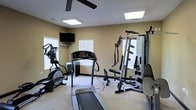 This single-floor fitness space is thoughtfully designed to support a comprehensive workout routine. The room features a neutral, light-toned color palette with beige walls and a white ceiling, creating a clean and focused environment. Natural light flows in through multiple windows fitted with white horizontal blinds, complemented by recessed flat-panel ceiling lights that ensure the space remains well-lit at all times. A ceiling fan adds comfort during intense workout sessions. The room is outfitted with a range of equipment, including a treadmill, an elliptical cross-trainer, and a multi-station cable and weight machine equipped with a pull-down bar and adjustable weight stack. A wall-mounted flat-screen television is positioned for easy viewing during cardio sessions, adding an element of entertainment. The flooring appears to be a light-toned carpet, providing a softer surface underfoot. The layout is practical and spacious enough to accommodate multiple users and exercise types simultaneously, making this fitness room a functional and self-contained space well-suited for daily training needs.