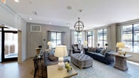 This refined single-floor space exudes classic elegance through its thoughtful design and carefully curated furnishings. The main lounge area is anchored by a striking orb chandelier that draws the eye upward to crisp white ceilings adorned with crown molding and recessed lighting. Large, floor-to-ceiling windows framed by sheer white drapes flood the interior with generous natural light, creating an open and airy atmosphere throughout.

The seating arrangement is both stylish and functional, featuring a pair of deep navy blue sofas, a tufted grey velvet ottoman serving as a central coffee table, and upholstered accent chairs in complementary tones. Decorative throw pillows with geometric and floral patterns add visual texture, while a soft blue area rug grounds the entire arrangement and adds warmth underfoot.

Console tables and side tables are tastefully styled with ceramic table lamps, greenery, and decorative accessories, contributing to the space's polished aesthetic. The walls are finished in a soft grey tone, providing a sophisticated neutral backdrop that ties the furnishings together harmoniously. The overall ambiance is one of understated luxury — welcoming, well-lit, and meticulously designed for both comfort and visual appeal.