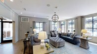 This refined single-floor space exudes classic elegance through its thoughtful design and carefully curated furnishings. The main lounge area is anchored by a striking orb chandelier that draws the eye upward to crisp white ceilings adorned with crown molding and recessed lighting. Large, floor-to-ceiling windows framed by sheer white drapes flood the interior with generous natural light, creating an open and airy atmosphere throughout.

The seating arrangement is both stylish and functional, featuring a pair of deep navy blue sofas, a tufted grey velvet ottoman serving as a central coffee table, and upholstered accent chairs in complementary tones. Decorative throw pillows with geometric and floral patterns add visual texture, while a soft blue area rug grounds the entire arrangement and adds warmth underfoot.

Console tables and side tables are tastefully styled with ceramic table lamps, greenery, and decorative accessories, contributing to the space's polished aesthetic. The walls are finished in a soft grey tone, providing a sophisticated neutral backdrop that ties the furnishings together harmoniously. The overall ambiance is one of understated luxury — welcoming, well-lit, and meticulously designed for both comfort and visual appeal.