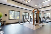 The Cahaba Grandview Fitness Room in Birmingham, Alabama offers a thoughtfully designed workout facility that balances function with a polished aesthetic. The space features a tray ceiling with crown molding detail, ceiling fans, and recessed fluorescent lighting that keep the environment bright and comfortable during any workout. Large, dark-framed windows line the walls, flooding the room with natural light and offering views of the surrounding greenery outdoors.

The flooring transitions between durable rubber gym mats and smooth concrete, providing appropriate surfaces for both equipment use and open movement. A striking orange-and-black cable crossover machine serves as a centerpiece, complemented by an array of strength training benches and selectorized weight machines positioned throughout the space. The cardio zone is anchored by Matrix-brand treadmills and a stair climber, catering to a full range of aerobic workouts.

A wall-mounted flat-screen television keeps members entertained, while large mirrors on one wall enhance the sense of space and allow for form checks during exercise. The entry corridor connects seamlessly to the main gym floor, featuring decorative artwork on the walls that adds a refined, community-oriented touch. An adjacent room with distinctive wallcovering provides additional utility space. Overall, the fitness room presents a clean, modern, and well-appointed environment suited for a comprehensive workout routine.