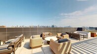 Located at 508 51st Street in West New York, New Jersey, this amenity floor at The Grand offers an impressive combination of fitness and outdoor living spaces. The hallway sets a polished tone with large-format tile flooring, exposed industrial-style ceilings with modern linear lighting, and a branded entry mat, accessed via a stainless steel elevator. The spacious exercise room features warm hardwood-style flooring, an exposed structural ceiling with sleek overhead lighting, and floor-to-ceiling glass walls that frame sweeping views of the New York City skyline. The fitness area is comprehensively equipped with cable machines, free weights, kettlebells, medicine balls, resistance equipment, and open floor space for functional training. Adjoining the gym is a generously sized rooftop terrace accessible through wide sliding glass doors, finished with large neutral-toned pavers and contemporary modular lounge seating. A second outdoor area showcases a striking green roof planted with low-growing sedum, alongside a pergola-shaded dining area and vibrant planter boxes, all set against a breathtaking panoramic backdrop of the Manhattan skyline. Together, these spaces offer a premium lifestyle amenity experience.
