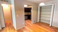This single-floor space offers a practical and comfortable layout with distinct living and kitchen areas. The kitchen is equipped with dark espresso-toned cabinetry that extends to the upper and lower sections, providing ample storage. Granite-style countertops in warm earth tones complement the dark wood finish, while a stainless steel double-basin sink and black electric range with coil burners complete the functional cooking space. A dishwasher and refrigerator are also present, making this a well-appointed kitchen. An arched doorway connects the kitchen to the adjacent dining or transitional area, which features warm hardwood flooring and a pendant light fixture overhead. Built-in open shelving is positioned nearby, offering convenient display or storage space. The living area is carpeted in neutral tones and benefits from a sliding glass door that floods the room with natural light and provides access to the outdoors. Neutral-toned walls throughout the space create a clean, versatile backdrop, allowing for a variety of interior styles. Overall, the space presents a straightforward, functional design suitable for comfortable everyday living.