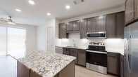 This single-floor residence showcases a thoughtfully designed interior with a consistent, contemporary aesthetic throughout. The kitchen serves as the focal point of the space, anchored by a generously sized center island topped with striking white-and-grey veined granite. Dark espresso-toned cabinetry lines the walls, complemented by a classic white subway tile backsplash with under-cabinet lighting. A full suite of stainless steel appliances — including a French door refrigerator with ice maker, electric range, over-the-range microwave, and dishwasher — equips the kitchen for practical everyday use. Light wood-look flooring runs throughout the main living and kitchen areas, creating a clean, cohesive look. The open layout allows sightlines from the kitchen island toward multiple doorways, reflecting a functional flow between spaces. The bathroom continues the design language with matching dark cabinetry, granite countertops, a framed mirror, and a brushed nickel vanity light fixture. The bedroom offers plush carpeting underfoot, a ceiling fan with integrated lighting, and wide sliding glass doors that invite natural light and provide access to an outdoor area.