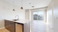 This single-floor residence showcases a contemporary design with a neutral palette of warm beige and white tones throughout. The open-concept layout connects the kitchen and living area seamlessly, centered around a sleek peninsula with a white quartz countertop, an undermount sink, and dark wood cabinetry. Pendant lighting with muted bronze fixtures hangs above the peninsula, complementing the stainless steel appliances, including a refrigerator, microwave, and range. Light hardwood flooring extends throughout the main living areas, adding warmth and continuity.

The living area is bathed in natural light through a large sliding glass door that opens onto a private balcony, offering views of the surrounding urban landscape. The two bedrooms feature plush neutral carpeting and large windows that bring in generous daylight while framing city views. The bathroom is finished with light hardwood flooring, a wood-toned vanity with a white countertop, and a tiled shower-tub combination. Clean lines, recessed and flush-mount lighting, and consistent finishes throughout contribute to a cohesive, modern aesthetic well-suited for contemporary living.