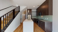 This two-story space showcases a cohesive contemporary design throughout, combining clean lines, quality finishes, and a neutral palette. The main level centers on a well-appointed kitchen, which serves as a focal point of the living area. Dark espresso-toned cabinetry extends nearly to the ceiling, complemented by open shelving, a sage-green subway tile backsplash, and stainless steel appliances including a range and microwave. A generous white waterfall island provides additional workspace and features an undermount sink with a modern arc faucet, with pendant lighting above. Light hardwood flooring runs throughout both levels, adding warmth to the overall aesthetic.

A sleek staircase with dark wood detailing and metal balustrades connects the floors. The upper level offers open, light-filled rooms with large windows that bring in natural light and views of the surrounding neighborhood. The upper landing is spacious, with the dark stair railing providing a consistent design element. A bathroom on the lower level features terrazzo flooring, a wood-toned vanity with an undermount sink, and clean white walls, delivering a refined, minimalist finish throughout the space.