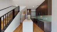 This two-story space showcases a cohesive contemporary design throughout, combining clean lines, quality finishes, and a neutral palette. The main level centers on a well-appointed kitchen, which serves as a focal point of the living area. Dark espresso-toned cabinetry extends nearly to the ceiling, complemented by open shelving, a sage-green subway tile backsplash, and stainless steel appliances including a range and microwave. A generous white waterfall island provides additional workspace and features an undermount sink with a modern arc faucet, with pendant lighting above. Light hardwood flooring runs throughout both levels, adding warmth to the overall aesthetic.

A sleek staircase with dark wood detailing and metal balustrades connects the floors. The upper level offers open, light-filled rooms with large windows that bring in natural light and views of the surrounding neighborhood. The upper landing is spacious, with the dark stair railing providing a consistent design element. A bathroom on the lower level features terrazzo flooring, a wood-toned vanity with an undermount sink, and clean white walls, delivering a refined, minimalist finish throughout the space.