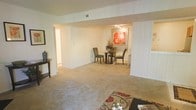 This single-floor residence offers an open and flowing layout that connects the living and dining areas in a cohesive, comfortable arrangement. The living area features plush wall-to-wall carpeting in a warm beige tone, creating a soft and inviting foundation throughout the space. A decorative area rug with red and black motifs anchors the living zone, adding a touch of visual interest to the neutral palette.

A structural beam and partial wall define the transition between the living area and the dining space beyond, maintaining an open feel while subtly distinguishing the two zones. The dining area accommodates a round glass-top table with upholstered chairs, positioned beneath track lighting that casts a warm, focused glow. A bold floral artwork in red and orange tones serves as a vibrant focal point on the dining room wall, providing contrast against the crisp white surroundings.

The walls throughout are painted in a clean, neutral white, enhancing the sense of brightness and space. A small console table in the living area adds a functional accent. Hallways visible on either side suggest additional rooms beyond, contributing to the overall practicality and accessibility of the layout.