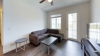 This single-floor unit presents a clean, modern aesthetic carried consistently throughout each room. The living area is an inviting space anchored by a large sectional sofa and coffee table, with wood-look flooring underfoot and a ceiling fan overhead. Natural light enters through a sizable window, while a glass-paneled door provides direct access to a private balcony, further brightening the space.

The kitchen is well-appointed, featuring a U-shaped layout with dark lower cabinetry contrasted by crisp white upper cabinets. Granite countertops extend across the workspace, complemented by stainless steel appliances including a range with microwave above. A granite-topped breakfast bar with bar seating connects the kitchen to the adjacent living area, supported by pendant lighting above.

The bedrooms are simply furnished with bed frames and mattresses, dark wood dressers, and nightstands. Each room includes horizontal blinds over windows that allow natural light, with wood-look flooring continuing throughout.

The bathrooms are finished with consistent granite vanity countertops, wood-look flooring, and a white tiled tub-shower combination. An additional half-bath provides added convenience, maintaining the unit's clean, neutral palette throughout.