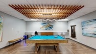 This well-appointed game room in Calgary, AB, offers a thoughtfully designed recreational space that balances energetic activity areas with stylish, contemporary interiors. The centerpiece of the room is a classic pool table with a vivid turquoise felt surface, positioned beneath a dramatic wood-lattice ceiling installation that adds warmth and architectural interest to the space. Sleek linear pendant lighting integrates seamlessly into the lattice structure, illuminating the area with a modern edge. Herringbone-patterned hardwood flooring runs throughout, adding a refined texture that complements the room's overall aesthetic. An air hockey table occupies one corner, flanked by vibrant large-scale canvas paintings featuring colorful depictions of animals, while a foosball table anchors another zone alongside a wall-mounted flat-screen television and a cozy lounge chair. Abstract artwork adorns the walls throughout, contributing to a lively and creative atmosphere. The entry features floor-to-ceiling glass walls and doors, flooding the space with light and providing an open, welcoming feel. A compact refreshment area with a mini-fridge is also present. The room is well-lit with recessed ceiling panels, ensuring a bright and inviting environment suitable for social recreation and leisure.