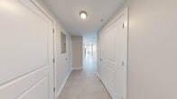 This single-floor unit features a distinctive industrial loft aesthetic, blending raw architectural elements with clean, modern finishes throughout. The space accommodates six bedrooms and one shared bathroom, all connected by a central hallway finished with light neutral-toned walls and large-format tile flooring that runs consistently throughout the unit.

The hallway serves as the main artery of the layout, lined with crisp white panel doors providing access to each bedroom. An electrical panel is accessible from the corridor, and ceiling-mounted light fixtures provide practical illumination. The bedrooms are modestly sized, with the primary bedroom offering slightly more space, and several rooms feature walk-in closets equipped with wire shelving systems.

The combined kitchen and living area is a standout space, featuring soaring exposed wood beam ceilings, visible metal ductwork, and a concrete structural column—hallmarks of adaptive industrial architecture. Large multi-pane windows flood this area with natural light while framing expansive urban rooftop views. The kitchen includes warm-toned wood cabinetry and stainless steel appliances.

The bathroom is cleanly appointed with tile flooring, a wood-vanity with white countertop, and a full soaking tub with subway-tiled surround and chrome fixtures.