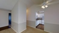 This single-floor residence offers a clean and functional layout designed for comfortable everyday living. The living area features soft carpeted flooring and is finished with neutral gray-toned walls accented by a chair rail detail, giving the space a classic, polished look. Vaulted ceilings rise overhead, creating an airy sense of openness, while a multi-light ceiling fan provides both style and practical air circulation. An open archway connects the living area to the kitchen, creating a seamless flow between spaces. The kitchen is well-appointed with white cabinetry that extends to the upper walls, offering generous storage. Contrasting dark countertops provide a visual anchor, complemented by white appliances including a range and over-the-range microwave. The kitchen floor features rich, dark hardwood-style flooring that stands in pleasing contrast to the carpeted living spaces. Recessed and under-cabinet lighting in the kitchen ensures a well-lit work environment. The overall ambiance of the space is bright, neutral, and inviting, with a consistent design palette that ties each area together cohesively.