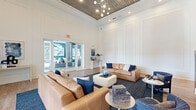 This single-floor space offers a refined and contemporary design across a generous footprint, combining open-plan living with distinct functional areas. The centerpiece is a dramatic living room boasting soaring double-height ceilings clad in warm wood paneling, a statement chandelier with globe pendants, and crisp white shiplap walls that amplify the sense of openness. Tan leather sofas, navy accent pillows, and a plush area rug create an inviting seating arrangement, while a sleek linear fireplace with an integrated wall-mounted display adds a striking focal point. Large glass doors flood the space with natural light and provide views of the outdoor surroundings. The kitchen is equally impressive, featuring a bold blue accent wall, warm wood cabinetry, marble-look countertops, stainless steel appliances, and a generously sized island with seating for multiple guests, all illuminated by stylish pendant lighting. Light hardwood-style flooring flows seamlessly throughout. The spacious primary bedroom offers a comfortable and private retreat. A dedicated conference or meeting room rounds out the layout, complete with a marble-top table, upholstered blue chairs, a whiteboard wall, and curated artwork, making it ideal for focused gatherings or collaborative work.