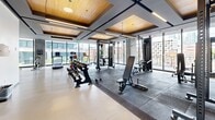 Located at 235 Rue Young in Montréal, this well-appointed fitness facility spans a generous single floor, offering a comprehensive and thoughtfully organized exercise environment. The gym is divided into distinct functional zones that cater to a wide range of training disciplines. Floor-to-ceiling windows line much of the perimeter, flooding the space with abundant natural light and providing open views of the surrounding Montréal cityscape, creating an energizing atmosphere throughout.

The cardio zone features treadmills, stationary spin bikes, and rowing machines arranged along the windowed walls to maximize natural light exposure. A dedicated strength and free weights area is equipped with adjustable benches, dumbbell racks, cable machines, and barbell stations, all set on rubber-matted flooring for safety and stability. Full-length mirrors are strategically positioned throughout to support proper form and enhance the sense of space.

Architecturally, the facility stands out with a refined ceiling design incorporating warm wood-paneled insets and recessed linear lighting, lending a polished, upscale aesthetic. The light-toned flooring contrasts elegantly with the darker rubber workout mats. Touches of greenery add life to the environment. The overall ambiance is clean, bright, and professional, making this fitness facility a premium destination for residents and users of 235 Rue Young.