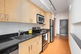 This thoughtfully designed single-floor residence combines modern aesthetics with functional living spaces. The galley-style kitchen is a standout feature, showcasing light wood cabinetry with brushed nickel hardware, dark granite countertops, a stainless steel sink, and a full suite of stainless steel appliances including a refrigerator, electric range, built-in microwave, and dishwasher. Warm bamboo-style hardwood flooring runs the length of the kitchen and entry corridor, illuminated by a sleek track lighting system overhead. The living area is generously sized, bathed in natural light from large floor-to-ceiling windows and sliding glass doors that open onto a private balcony with outdoor views. Soft, neutral-toned carpeting grounds the space, creating a comfortable and inviting atmosphere. The bedroom offers a similarly bright and open feel, with large windows, plush carpeting, and a double sliding door closet finished in the same warm wood tones found throughout the unit. The bathroom is finished with matching dark granite countertops, light wood vanity cabinetry, a vessel-style sink, and a shower-tub combination. Clean white walls and consistent warm wood accents tie the entire space together with a cohesive, contemporary aesthetic.
