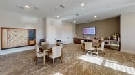 This single-floor space is thoughtfully designed as a multifunctional gathering and activity area, combining comfort with practicality. The layout accommodates several distinct seating zones, each furnished with upholstered chairs and dark wood tables, creating an inviting environment for group activities or casual meetings. Natural light streams in through large windows fitted with horizontal blinds and sheer curtains, brightening the neutral-toned walls and patterned carpet flooring throughout.

One area functions as a lounge near the entry, featuring armchairs, an ottoman, and decorative accessories alongside views of the surrounding exterior. Adjacent to this is a more intimate dining or game area with a round table and patterned upholstered chairs beneath a warm pendant light and coordinating wall art.

The main activity room is generously sized, accommodating multiple rectangular tables and chairs alongside a wall-mounted flat-screen display and a credenza with storage. A chalkboard on one side adds an educational element. Recessed lighting and ceiling speakers enhance functionality throughout. Thoughtful touches including framed artwork, indoor greenery, and coordinated furnishings contribute to a warm, welcoming ambiance well-suited for community programming and collaborative use.