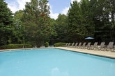 Located at 3400 Peachtree Corners Circle in Peachtree Corners, Georgia, this single-level outdoor amenity area offers a well-appointed community pool and recreation space designed for relaxation and leisure. The centerpiece of the space is a large, rectangular swimming pool with clear blue water, surrounded by an expansive concrete sundeck that provides ample room for lounging and movement. A generous arrangement of reclining lounge chairs lines the perimeter of the deck, offering multiple options for sunbathing or unwinding poolside. Several shaded seating areas are positioned throughout the patio, each featuring round metal tables with coordinating chairs and teal market umbrellas, ideal for outdoor dining or social gatherings. Lush, mature landscaping—including tall pine trees and manicured shrubbery—frames the space on all sides, creating a private, natural atmosphere that feels secluded from the surrounding area. A low iron fence defines the boundary of the pool enclosure, while a clubhouse-style building with a pitched roof and large windows is situated adjacent to the deck, likely providing interior amenity access. The overall ambiance is serene and park-like, with the dense Georgia tree canopy contributing a sense of shade, privacy, and natural beauty throughout the space.