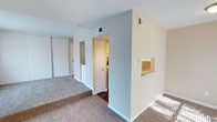 This single-floor space is finished in a clean, neutral palette, with light beige and soft gray walls complemented by white trim throughout. The main living area features wall-to-wall carpet in a warm tan tone, lending a comfortable, understated feel underfoot. A structural half-wall with a built-in pass-through window serves as a functional divider between the living area and an adjacent space, providing both visual interest and practical connectivity between rooms.

Along the back wall of the main area, a sliding mirrored closet door reflects natural light and adds depth to the space. A compact built-in dresser or storage unit with white drawer fronts is positioned nearby, offering convenient built-in organization. Ceiling-mounted HVAC vents are distributed throughout, ensuring efficient climate control across the layout.

A doorway leads into a secondary room that transitions to hardwood-style flooring, providing a clear distinction between the two areas. White interior doors and consistent trim work unify the overall design. Natural light filters in, brightening the space and highlighting the clean, move-in-ready condition of the interior. The layout flows efficiently, making practical use of the available footprint.