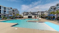 Located at 2700 Whakatane Ln in Fuquay-Varina, NC, this two-floor residential community showcases a striking craftsman-inspired architectural design, blending board-and-batten siding, stone accents, and dark pitched rooflines across its multi-building layout. The exterior palette of slate blue and gray tones lends the community a cohesive, upscale aesthetic.

At the heart of the community is an expansive resort-style aquatic area featuring multiple swimming pools with crystal-clear water, zero-entry points, and dedicated lap sections. Surrounding the pools is a generously sized sundeck furnished with lounge chairs and shaded umbrella stations, providing ample space for relaxation. A well-appointed pool house with large glass doors serves as the central amenity hub, offering direct access to the water and additional facilities within.

Residents on upper floors enjoy private balconies overlooking the pool courtyard, while ground-level units open to landscaped surroundings featuring palm trees and lush greenery that enhance the resort atmosphere. A separate spa or wading pool area adds to the layered aquatic offerings, complemented by pergola-shaded seating zones.

The overall ambiance of the outdoor space is bright, open, and inviting, with abundant natural light and an open sky setting that reinforces the community's resort-inspired character throughout.