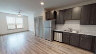 This single-floor residence is thoughtfully designed with a contemporary aesthetic, combining functionality and clean, modern finishes. The open-concept layout connects the kitchen and living area seamlessly, creating a spacious and versatile environment. Light wood-look flooring extends throughout the main living spaces, complementing the neutral color palette and lending warmth to the interior.

The kitchen is well-appointed with dark espresso-toned cabinetry that reaches toward the ceiling, providing ample storage above and below. Light marbled laminate countertops contrast attractively against the cabinetry, and a full suite of stainless steel appliances — including a refrigerator, dishwasher, electric range, and built-in microwave — ensures practical daily functionality. A double-basin sink with a modern faucet completes the kitchen workspace.

The bedroom features plush neutral carpeting, a ceiling fan, and a window fitted with blinds that allows natural light while maintaining privacy. The bathroom presents a long vanity with dark cabinetry and a marbled countertop, a wide mirror with overhead lighting, wood-look flooring, and a bathtub. A louvered double-door closet is conveniently positioned near the living area, adding practical storage to the unit.