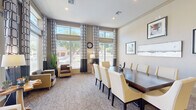 This single-floor community space is designed to accommodate both collaborative gatherings and individual productivity. The main area is anchored by a large, dark-wood conference table surrounded by cream and yellow upholstered chairs, providing ample seating for meetings or group events. Patterned carpeting with a geometric design covers the floors throughout, adding visual interest and a polished aesthetic.

Floor-to-ceiling windows line one wall, dressed with bold black-and-white patterned drapes and roman shades, flooding the space with natural light and offering views of the surrounding outdoor greenery. The elevated ceiling height enhances the open, airy feel of the room. A lounge area with cushioned armchairs is positioned near the windows, creating a comfortable spot for informal conversation.

A dedicated workstation area features a long granite-topped desk with dual computer setups, mesh office chairs, built-in shelving, and framed artwork above — making it a functional space for business tasks. Wall sconces and recessed lighting provide layered illumination throughout. Curated artwork displayed on neutral-toned walls and decorative plants contribute to a refined, welcoming ambiance.