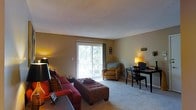 This single-floor space showcases a thoughtfully arranged open-concept layout that seamlessly combines the living and dining areas into one cohesive environment. The living area is anchored by plush carpeting underfoot and warm, tan-toned walls that create an inviting and comfortable atmosphere. A large sliding glass door dominates the far wall, flooding the space with natural light and offering views of surrounding greenery, while also providing direct access to an outdoor balcony enclosed by a railing — ideal for enjoying fresh air. The living area comfortably accommodates a full seating arrangement, including a sofa and an accent armchair, complemented by a tufted ottoman that adds both style and function. Adjacent to the living area, the dining space features a dark-toned table and chair set, illuminated by a warm table lamp that adds a cozy ambiance to the corner. The smooth, light-colored ceiling enhances the sense of openness throughout the room. Overall, this space balances practicality and comfort, offering a welcoming environment with natural light, outdoor connectivity, and a well-defined yet open living arrangement.