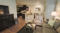 This single-floor space showcases an open-concept layout that connects the living, dining, and kitchen areas into one cohesive, flowing environment. The warm hardwood flooring extends throughout, providing a sense of continuity and grounding the space with rich, natural tones.

The living area is anchored by a dark espresso coffee table and complemented by two cream upholstered armchairs with nailhead trim detailing, accented with chevron-patterned throw pillows. A patterned area rug defines the seating zone and adds texture to the overall design.

The kitchen features dark espresso cabinetry contrasted against light countertops, with an island or peninsula providing both additional workspace and a natural boundary between cooking and living spaces. Pendant lighting hangs above, adding warmth and a polished finishing touch. Full-size appliances, including a black refrigerator and microwave, complete the functional kitchen setup.

Adjacent to the kitchen, the dining area accommodates a table and chairs beneath natural light from a nearby window. A framed mirror on the wall enhances the sense of openness. Track lighting on the ceiling provides flexible illumination throughout, contributing to the space's bright, welcoming ambiance.