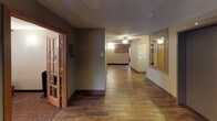 This single-floor building offers a thoughtfully designed layout centered around functional common areas and easy navigation. The main entry features a glass-paneled door that allows natural light to filter into the foyer, which is equipped with elevator access and a welcoming entry mat. Warm wood-look flooring runs throughout the corridors, complemented by dark baseboards and neutral beige and taupe wall tones that create a cohesive, understated aesthetic.

The hallways are well-lit with a combination of ceiling-mounted fixtures and wall sconces, maintaining a comfortable ambiance throughout. Ceiling fans with light kits are present in key areas, contributing to both ventilation and illumination. Wood-trimmed interior doors with warm honey-toned finishes add a consistent design element across the space.

A dedicated mailroom alcove houses a wall of individual mailboxes, providing a convenient centralized mail collection point for residents. Nearby, posted notices and informational boards serve as a community communication hub. A glimpse into an adjacent area reveals fitness equipment, indicating an on-site exercise space available to occupants. The layout connects all areas seamlessly, making this building's common spaces both practical and welcoming for everyday use.