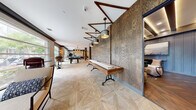 Located at 3964 Watercourse Dr in Fort Worth, TX, this clubhouse delivers an upscale, industrial-rustic atmosphere across a single, generously sized floor. The space blends warm wood tones, textured wall treatments, and modern fixtures throughout, creating a cohesive and inviting environment.

The main gathering area features distinctive geometric patterned flooring, a full-size pool table beneath a striking wagon-wheel chandelier, and large windows that flood the space with natural light while offering views of the surrounding greenery. A shuffleboard table runs alongside a richly textured wood accent wall, complemented by a series of angular pendant lights overhead.

A dedicated bar area serves as a central social hub, boasting an extended island with bar seating, exposed wooden ceiling beams, white subway tile walls, ornate pendant lighting, and dark granite countertops with built-in refrigeration. Adjacent to the bar is a cozy lounge nook outfitted with leather swivel chairs, a cowhide rug, dark shiplap walls, and a large-format industrial artwork — a nod to Fort Worth's heritage.

A media lounge area features a large wall-mounted flat-screen TV framed in reclaimed wood, with plush sectional seating and Texas-themed artwork. Throughout the clubhouse, the thoughtful layering of materials — wood, tile, metal, and leather — creates a refined yet welcoming space ideal for social gatherings.