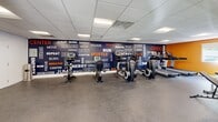 Located at 2900 Knights Rd in Bensalem, PA, this fitness center offers a well-equipped, single-floor exercise space designed to support a full range of workout routines. The room features a drop-panel ceiling with recessed fluorescent lighting that ensures bright, even illumination throughout. The flooring is a durable rubber material suited for high-impact activity, transitioning to carpet in the entry area.

The exercise room is divided into distinct functional zones. A dedicated cardio section lines one wall and features stationary bikes and treadmills positioned in front of a bold, floor-to-ceiling motivational mural in navy blue and orange, displaying energizing words such as "Run," "Hustle," "Energy," and "Grind." A strength training area offers a multi-station cable machine, adjustable weight benches, and a tiered dumbbell rack with a full-length mirror backdrop, allowing users to monitor their form. Natural light enters through windows, offering views of the surrounding greenery.

The entry vestibule provides a clean, welcoming transition into the facility, complete with wall-mounted coat hooks, a large mirror, and a fire safety station. The overall design is functional and motivating, combining energetic aesthetics with practical equipment placement to create an accessible and comprehensive fitness environment.
