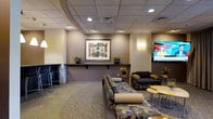 This single-floor lounge is thoughtfully designed to provide a comfortable and inviting environment for visitors and guests. The space features a drop tile ceiling with recessed lighting that casts a warm, even glow throughout the room, complemented by wall-mounted sconces that add an additional layer of ambient light.

The seating area is anchored by a large, patterned sofa and coordinating armchairs, offering ample space for relaxation or casual conversation. A cluster of round side tables provides convenient surfaces for guests, while a wall-mounted flat-screen television serves as a focal point on one end of the room. On the opposite side, a raised bar-height counter with sleek black stools offers an alternative seating option, ideal for those who prefer an elevated perch.

A decorative ledge runs along one wall, displaying framed abstract artwork and lush potted plants, lending the space a curated, refined aesthetic. The neutral warm-toned walls and carpeted flooring tie the design together cohesively, creating a relaxed yet polished atmosphere. Overall, this lounge strikes a balance between functionality and comfort, making it well-suited as a waiting area, social hub, or casual meeting space.