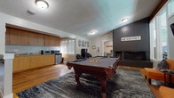 This single-story amenity space is designed to serve as a dynamic community gathering hub, blending recreational, social, and functional areas under one roof. The architectural design incorporates vaulted ceilings with an exposed wood beam, large picture windows, and transom-style glazing that flood the interior with abundant natural light while offering views of the surrounding greenery.

The central game room is the focal point of the space, anchored by a full-size pool table set atop a textured area rug over warm hardwood flooring. A dramatic dark brick fireplace with a raised hearth provides a bold visual statement, complemented by a wall-mounted flat-screen television and a pair of vibrant orange accent chairs for comfortable lounging. Playful signage, including "CAFE" and "NO VACANCY" lettering, adds a whimsical, eclectic character to the room.

Adjacent to the game room, a compact kitchenette is outfitted with warm wood cabinetry, granite-style countertops, a built-in sink, microwave, toaster oven, and a compact refrigerator — ideal for casual refreshments. A separate workstation nook features carpeted flooring, a dedicated computer desk, and a glass entry door that opens to an outdoor courtyard, bathing the area in soft natural light. A fitness area is also visible through interior windows, completing this well-rounded amenity space.