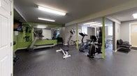 This fitness center offers a comprehensive workout environment designed to support a variety of exercise routines. The space is finished with durable speckled rubber flooring throughout, providing a safe and resilient surface ideal for both cardio and strength training activities. Vivid green accent walls contrast with neutral gray tones, creating an energizing atmosphere enhanced by bright fluorescent overhead lighting.

The cardio section is well-appointed with multiple stationary bikes, an elliptical trainer, and a treadmill, giving users a range of options for aerobic exercise. A wall-mounted flat-screen television is positioned for easy viewing during workouts. For strength training, a full dumbbell rack lines one wall, offering a wide selection of free weights to accommodate various fitness levels.

Large mirrors are strategically placed along the walls, visually expanding the space and allowing users to monitor their form. Structural columns, finished in green to match the accent walls, add a design cohesion throughout the room. A Dasani water vending machine is conveniently located within the facility, ensuring hydration is readily accessible. Additional doors provide utility access and storage. A decorative potted tree adds a touch of warmth to the otherwise functional space, rounding out this practical and well-organized fitness facility.