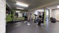 This fitness center offers a comprehensive workout environment designed to support a variety of exercise routines. The space is finished with durable speckled rubber flooring throughout, providing a safe and resilient surface ideal for both cardio and strength training activities. Vivid green accent walls contrast with neutral gray tones, creating an energizing atmosphere enhanced by bright fluorescent overhead lighting.

The cardio section is well-appointed with multiple stationary bikes, an elliptical trainer, and a treadmill, giving users a range of options for aerobic exercise. A wall-mounted flat-screen television is positioned for easy viewing during workouts. For strength training, a full dumbbell rack lines one wall, offering a wide selection of free weights to accommodate various fitness levels.

Large mirrors are strategically placed along the walls, visually expanding the space and allowing users to monitor their form. Structural columns, finished in green to match the accent walls, add a design cohesion throughout the room. A Dasani water vending machine is conveniently located within the facility, ensuring hydration is readily accessible. Additional doors provide utility access and storage. A decorative potted tree adds a touch of warmth to the otherwise functional space, rounding out this practical and well-organized fitness facility.