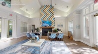 This single-story community clubhouse offers a thoughtfully designed layout across a generous footprint, seamlessly blending social, functional, and recreational spaces. The centerpiece is a soaring vaulted living area anchored by a striking floor-to-ceiling chevron-patterned accent wall in teal, gold, and natural wood tones, framing a fireplace with a mounted flat-screen TV above. Rich hardwood-style flooring, abundant natural light from large windows and French doors, and crisp white millwork — including elegant columns and crown molding — lend the space a bright, welcoming ambiance. A connected lounge and dining area provide flexible seating arrangements for residents to gather or work. The fitness center is well-equipped with cardio machines including treadmills and ellipticals, a multi-function cable machine, free weights, and a wall of mirrors, all beneath vaulted ceilings and large windows overlooking lush greenery. A separate lounge room features vibrant teal accent walls, plush seating, and a wall-mounted TV, creating a comfortable media or multipurpose space. The on-site laundry room offers multiple commercial-grade washers and dryers set against cheerful light-blue walls with hardwood-style flooring. Connected hallways tie the entire facility together efficiently.