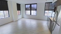 This single-floor space offers a clean, contemporary aesthetic defined by an open layout and an abundance of natural light. The room is anchored by smooth light-toned hardwood flooring that stretches across the entire area, lending warmth and a sense of spaciousness to the interior.

Large, multi-pane windows with sleek black frames dominate the walls, flooding the space with natural light while framing views of the surrounding urban streetscape. Each window is fitted with black roller blinds, providing practical light control and privacy. The windows' generous proportions and city-facing orientation give the space an elevated, connected feel with its surroundings.

The walls are finished in a crisp, clean white, contributing to the bright and airy ambiance throughout. The minimalist design ethos is consistent across the space — there are no unnecessary architectural embellishments, allowing the natural light, flooring, and window arrangement to serve as the defining features.

The open floor plan offers considerable flexibility, making the space well-suited for a variety of uses. Overall, the space presents a modern, light-filled environment with a neutral palette and urban character that provides a versatile and inviting setting.