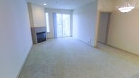 This single-floor space centers around a well-appointed living area that combines functional design with a warm, inviting ambiance. The room is finished with plush wall-to-wall carpeting in a neutral tone, providing a soft and comfortable foundation that complements the clean white walls throughout.

A standout feature of the living area is the tile-surround fireplace positioned along one wall, adding a classic focal point and a sense of warmth to the space. Adjacent to the fireplace, a side window draws in additional natural light, enhancing the overall brightness of the room.

Generous natural light fills the space through large sliding glass doors that open to an outdoor balcony, offering a seamless connection to the exterior and views of surrounding greenery. The sliding doors are dressed with vertical blinds, allowing for flexible light and privacy control.

A recessed ceiling light provides supplemental illumination, while a floor vent ensures efficient climate management. An open doorway visible toward the rear of the space hints at additional rooms beyond, suggesting a practical and connected layout. Overall, the space presents a clean, neutral palette well-suited for comfortable everyday living.