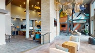 This expansive single-floor communal space makes a striking first impression with its soaring double-height ceilings, exposed steel trusses, and floor-to-ceiling windows that flood the interior with natural light and offer open views of the surrounding area. The architectural framework combines stacked-stone accent columns, polished tile flooring, and patterned carpeting to define distinct zones within the open layout.

The main lounge area features an eclectic mix of seating, including leather sofas, upholstered chairs, and wooden coffee tables arranged beneath oversized woven pendant lights and copper-toned hanging fixtures. A bold, large-scale pop-art mural dominates one section of the space, energizing a game and recreation area complete with a foosball table and wall-mounted televisions.

A dedicated dining and kitchenette zone showcases a shimmering blue tile accent wall, under-counter beverage refrigerators, dark countertops, and a rustic wood communal table with red accent chairs. Booth-style seating with tall wooden backs lines the window wall, providing semi-private spots bathed in natural light.

A workroom corridor leads to additional functional spaces, while a neon "Do What You Love" sign near the reception desk reinforces the space's energetic, community-oriented character throughout.
