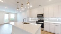Located at 1 Riverside Ave in Jacksonville, FL, this thoughtfully designed one-bedroom, one-bathroom unit offers a clean, modern aesthetic throughout. Light-toned hardwood-style flooring flows seamlessly across the entire space, complemented by crisp white walls and recessed lighting that keep the atmosphere bright and airy.

The open-concept kitchen and living area serves as the heart of the unit. The kitchen features a large white quartz island with an undermount sink, sleek white cabinetry reaching toward the ceiling, a soft grey tile backsplash, and stainless steel appliances including a range, microwave, and refrigerator. A cluster of globe pendant lights hangs above the island, adding a stylish focal point. A stacked washer and dryer is conveniently tucked into a dedicated closet just off the kitchen.

The living area extends toward a glass door and adjacent window that open to an outdoor balcony, filling the space with natural light and offering views of the surrounding Jacksonville skyline and greenery.

The bedroom is a comfortable, well-proportioned room with a ceiling fan, two windows framing city views, and access to the outdoors. A generous walk-in closet with wire shelving provides ample storage.

The bathroom is finished in a clean, contemporary style with a dual-sink vanity topped in white quartz, a large mirror with globe sconce lighting, and a glass-enclosed shower with white tile surround.