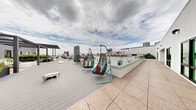 Located at 1509 Broadway in San Diego's vibrant downtown, this expansive rooftop amenity deck offers a well-appointed outdoor retreat with sweeping panoramic views of the city skyline. The deck is designed in a U-shaped layout, maximizing open-air space and creating distinct zones for relaxation and socializing.

A dedicated sun-lounging area features rows of sling chaise lounges set atop lush artificial turf, shaded by large, bold red cantilever umbrellas. Surrounding planters are filled with mature greenery and succulents, providing a garden-like atmosphere. Nearby, a built-in outdoor fireplace serves as a focal point for a cozy seating cluster with woven lounge chairs in teal and orange tones.

A separate open-air lounge area offers modular sofa seating, accent chairs, and side tables, all positioned to take full advantage of the dramatic San Diego skyline backdrop. Suspended egg-shaped swing chairs add a playful, modern element to the deck's aesthetic. Throughout the space, glass panel railings ensure unobstructed sightlines across the cityscape.

Wide-plank composite decking and large-format tile flooring lend a clean, contemporary finish underfoot. The modern white architectural facade of the building frames the rooftop, with floor-to-ceiling windows looking out onto the deck below. This rooftop space combines functional amenity design with resort-inspired style in the heart of downtown San Diego.