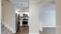 This two-story home offers a well-designed layout with modern finishes throughout. The kitchen is a functional centerpiece, featuring stainless steel appliances including a gas range, built-in microwave, and a full-size refrigerator. White shaker-style cabinetry pairs with a tile backsplash to create a clean, contemporary aesthetic. A track lighting fixture illuminates the space, complementing the textured ceiling. Crown molding adds a refined architectural detail that carries through the adjoining areas. Dark hardwood-style flooring grounds the space with warmth and contrast against the neutral, light beige walls. A staircase with white balusters and a wood handrail is visible from the main level, providing access to the second floor. The open sightlines between the kitchen and adjacent spaces contribute to an airy, connected feel throughout the main level. The overall design strikes a balance between practicality and style, with thoughtful details such as crown molding and quality finishes elevating the everyday living experience across both floors.