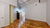 This urban loft-style space blends industrial character with modern finishes throughout its single-floor layout. The open living area immediately impresses with soaring ceilings supported by exposed white-painted beams, from which black iron pipes and pendant lighting run dramatically overhead. Rich, warm-toned hardwood floors extend throughout the space, adding timeless character and visual continuity.

The kitchen is thoughtfully designed with dark cabinetry, stainless steel appliances including a gas range, built-in microwave, and dishwasher, complemented by a white subway tile backsplash and open wooden shelving. A deep single-basin sink with a matte black faucet anchors the clean white countertops.

The bedroom features a continuation of the hardwood flooring, a window with an exterior brick view, and a painted brick accent wall that nods to the building's industrial heritage. Natural light filters in through black-framed windows, enhancing the airy feel of the room.

The bathroom is finished with large-format white wall tiles in the tub-shower surround, dark slate-toned floor tiles, a dark wood vanity with a white basin, matte black fixtures, and a round mirror — delivering a clean, contemporary aesthetic that ties the space together cohesively.